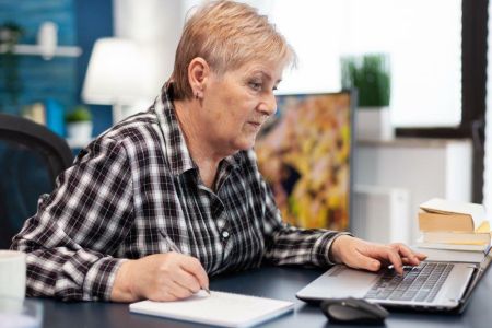 Starsza pani pisząca jedną ręką na laptopie a drugą notuje