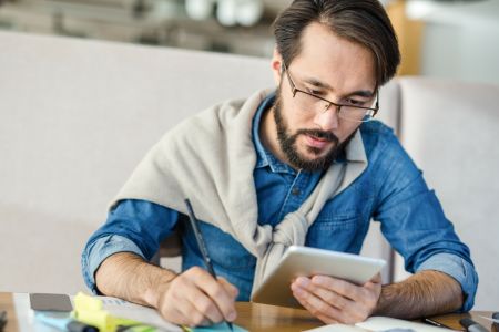 Mężczyzna siedzący przy biurku jedną ręką notujący coś na kartce a w drugiej trzymający tablet