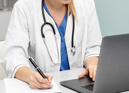 Kobieta doktor siedząca przy biurku w białym płaszczu lekarskim, przepisująca coś z laptopa