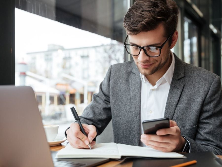 Mężczyzna siedzący przy biurku z laptopem i smartfonem w dłoni i notującym coś w zeszycie